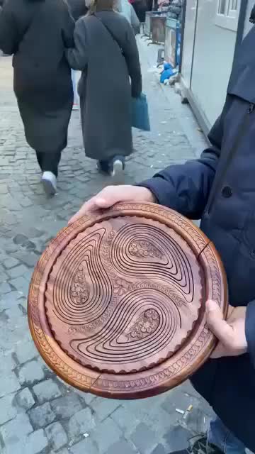 This may contain: a man is holding a wooden plate with designs on it in the middle of a cobblestone street