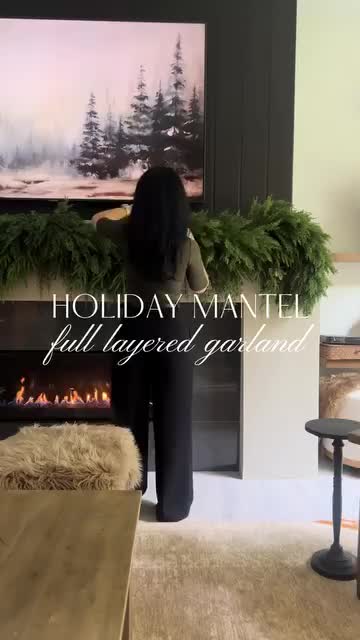 This may contain: a woman standing in front of a fireplace with a christmas wreath on top of it