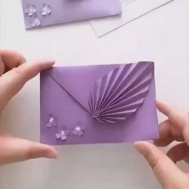 This may contain: two hands holding a purple envelope with paper flowers on the front and bottom, while another hand holds an envelope in the background