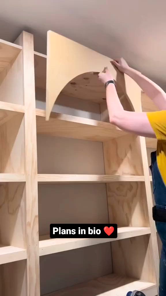 This may contain: a man in yellow shirt and blue jeans working on shelving unit with wooden shelves