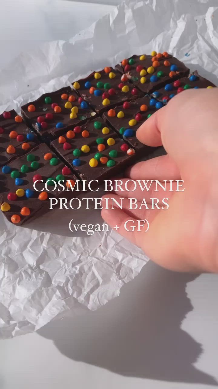 This may contain: a hand is holding a chocolate bar with colorful candy on it and the words cosmic brownie protein bars vegan gf