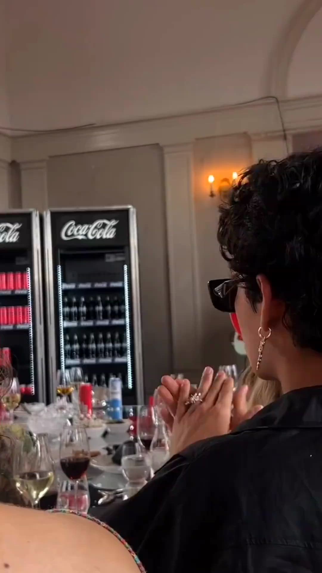 This may contain: a woman sitting at a bar with wine glasses in front of her and bottles behind her