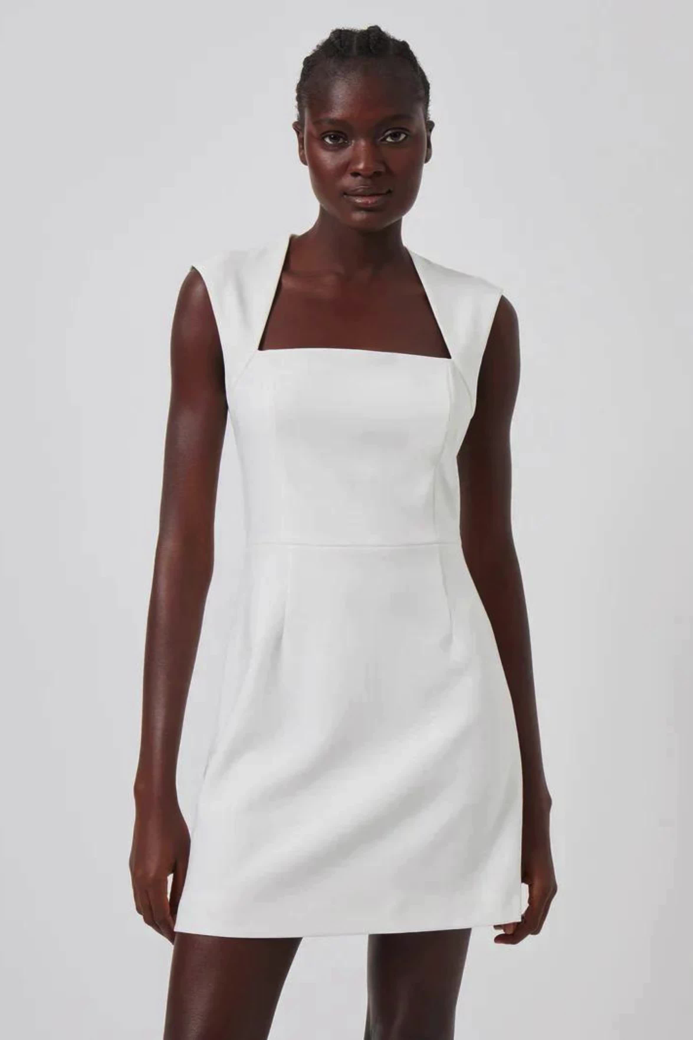 This contains: A front-facing view of a woman in a white, tailored mini dress with broad shoulder straps and a square neckline, posing in front of a white background.