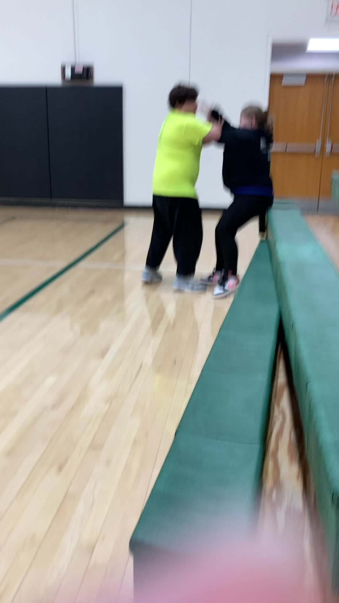 This may contain: two young children are dancing on a wooden floor in an indoor gym with hard wood floors and green benches