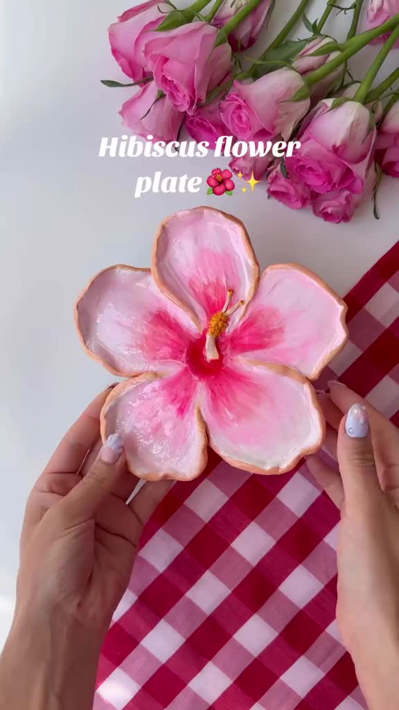 This may contain: a person holding a flower shaped cookie in front of pink flowers on a red and white checkered table cloth