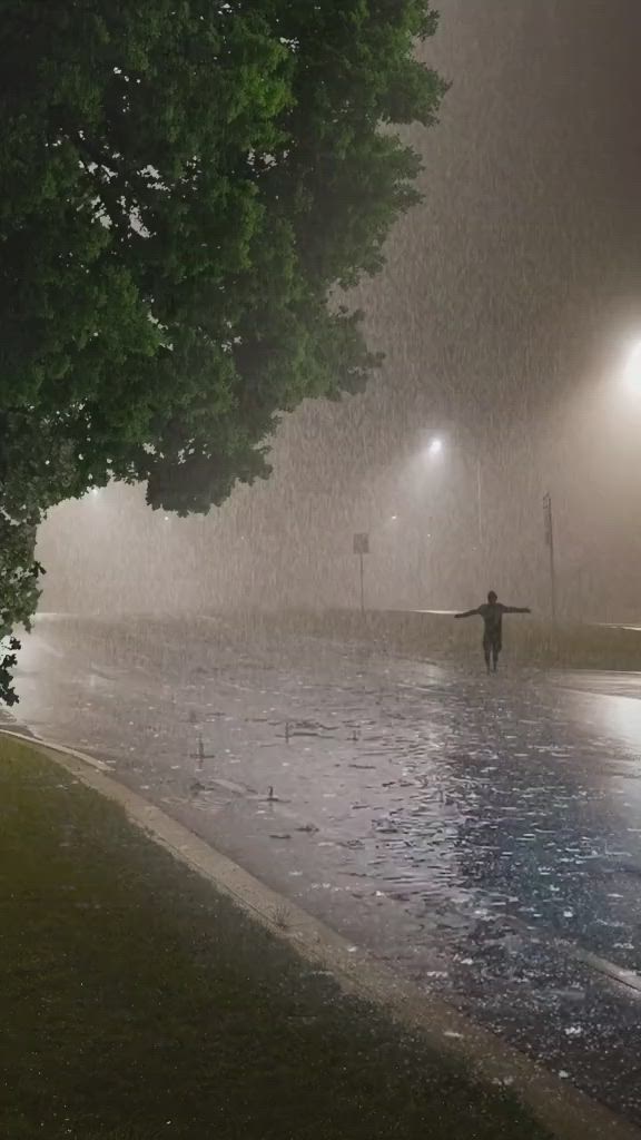 This may contain: a person standing in the rain at night with their arms spread out, holding an umbrella