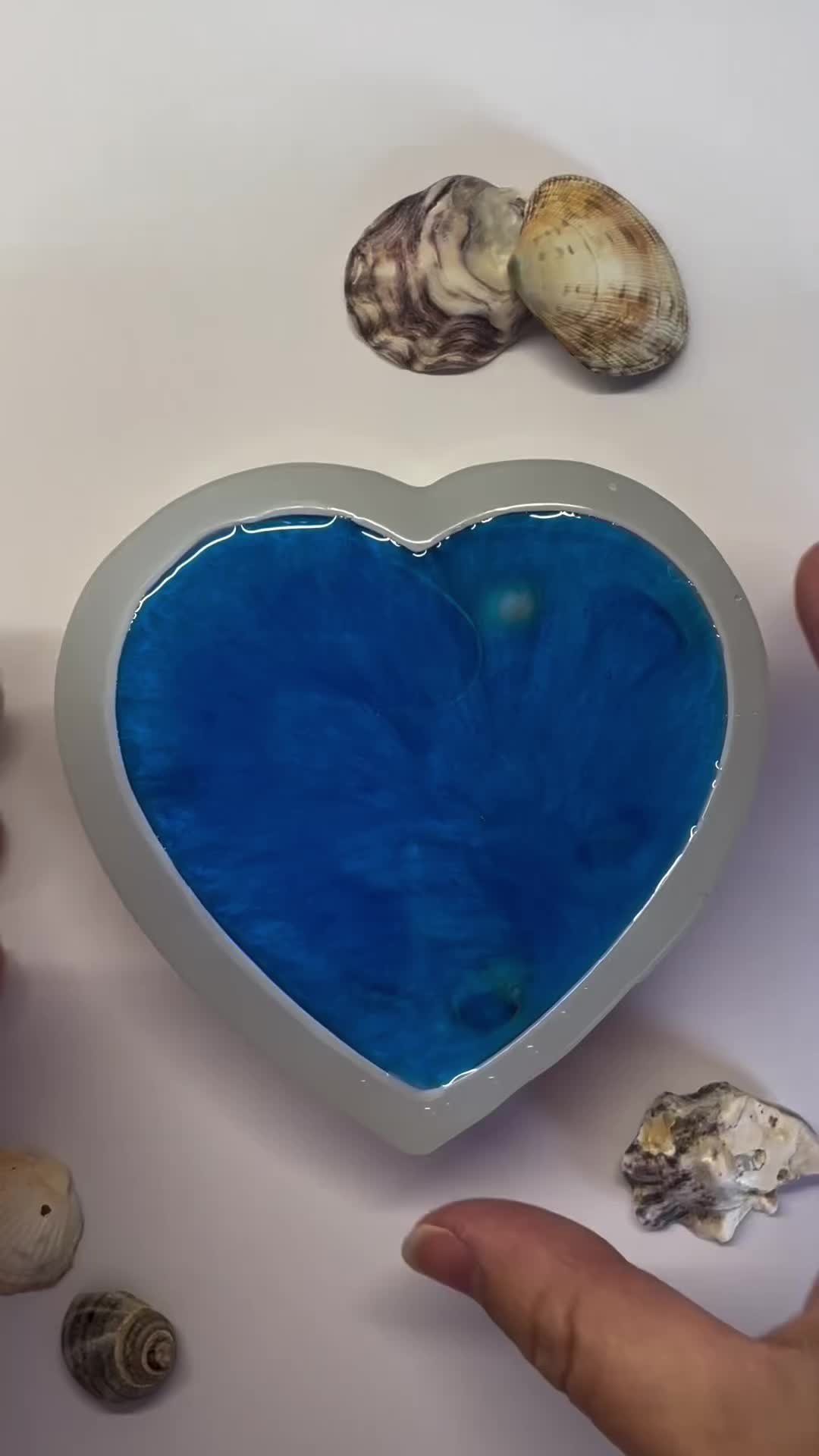 This may contain: a heart shaped bowl filled with blue liquid surrounded by sea shells