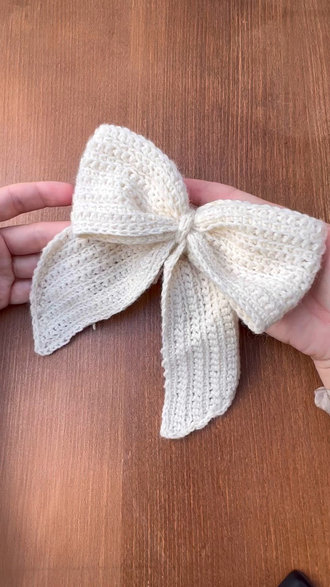 This may contain: a hand holding a white knitted bow on top of a wooden table