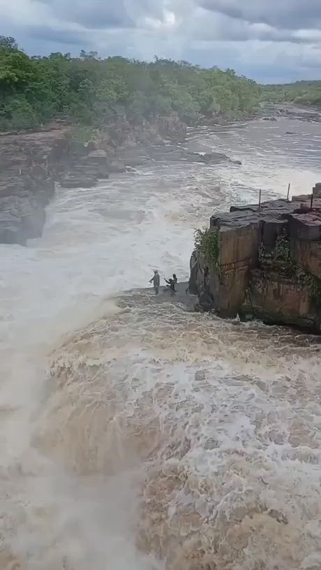 This may contain: two people standing on the edge of a cliff in water with waves crashing over it