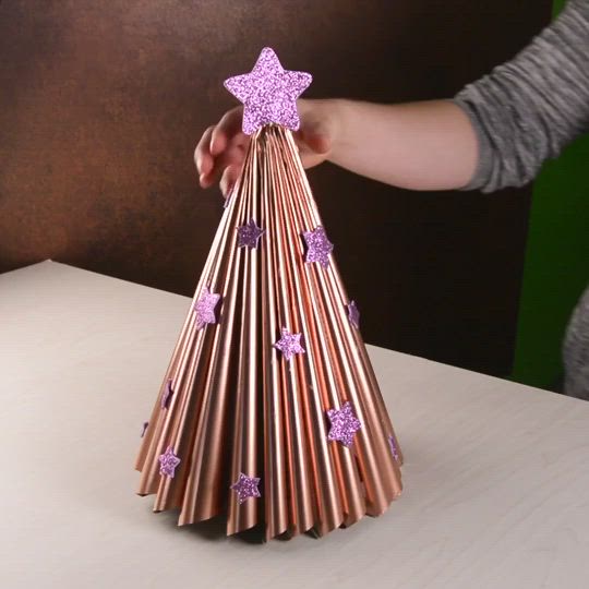 This may contain: a person is holding up a paper christmas tree made out of copper tubes and purple stars