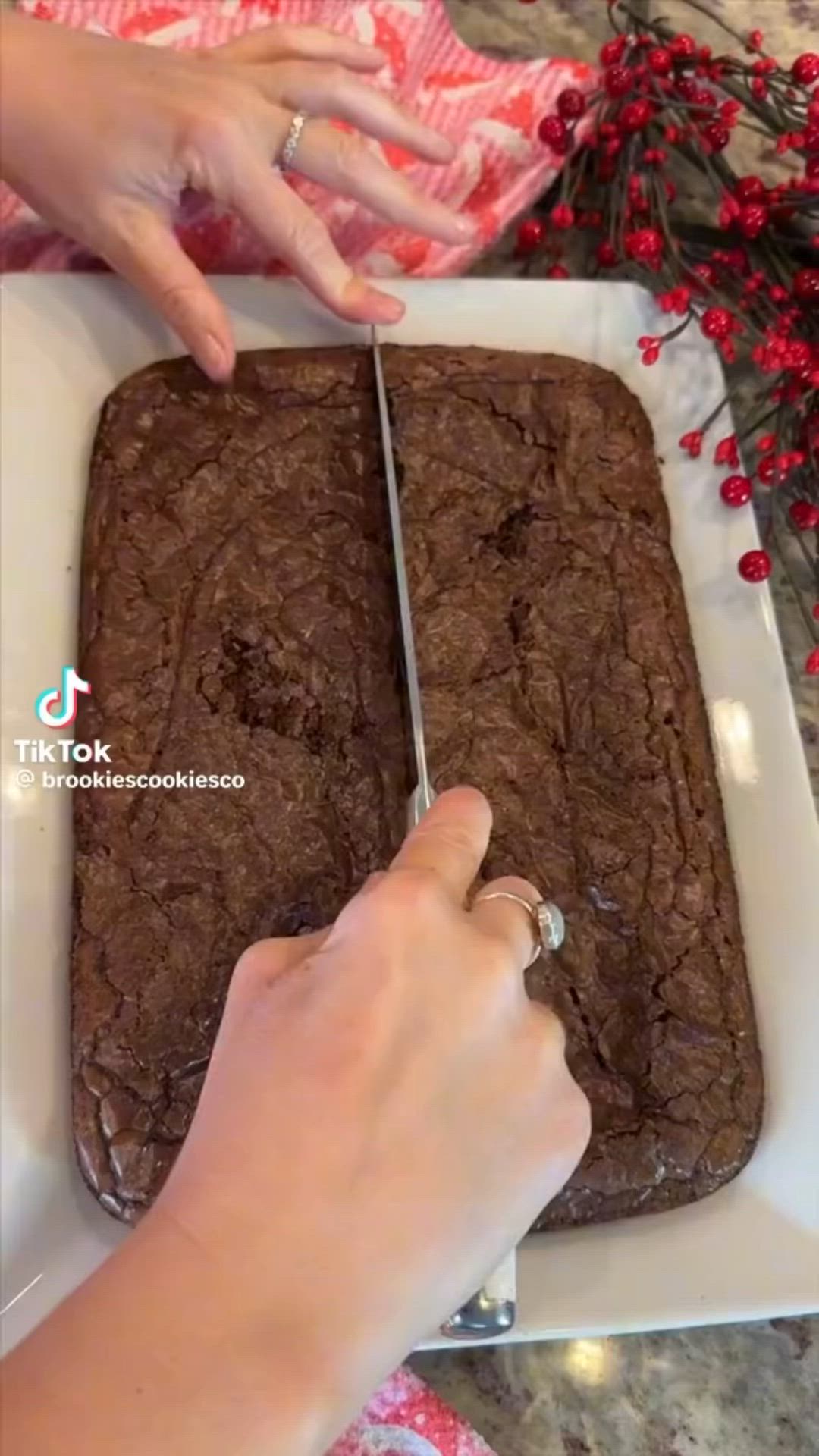 This may contain: a person cutting into a chocolate cake on a white platter with red flowers in the background