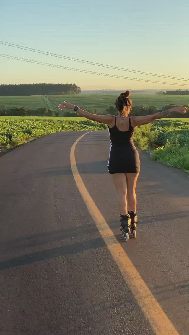 This may contain: a woman riding roller skates down a road next to a lush green field at sunset