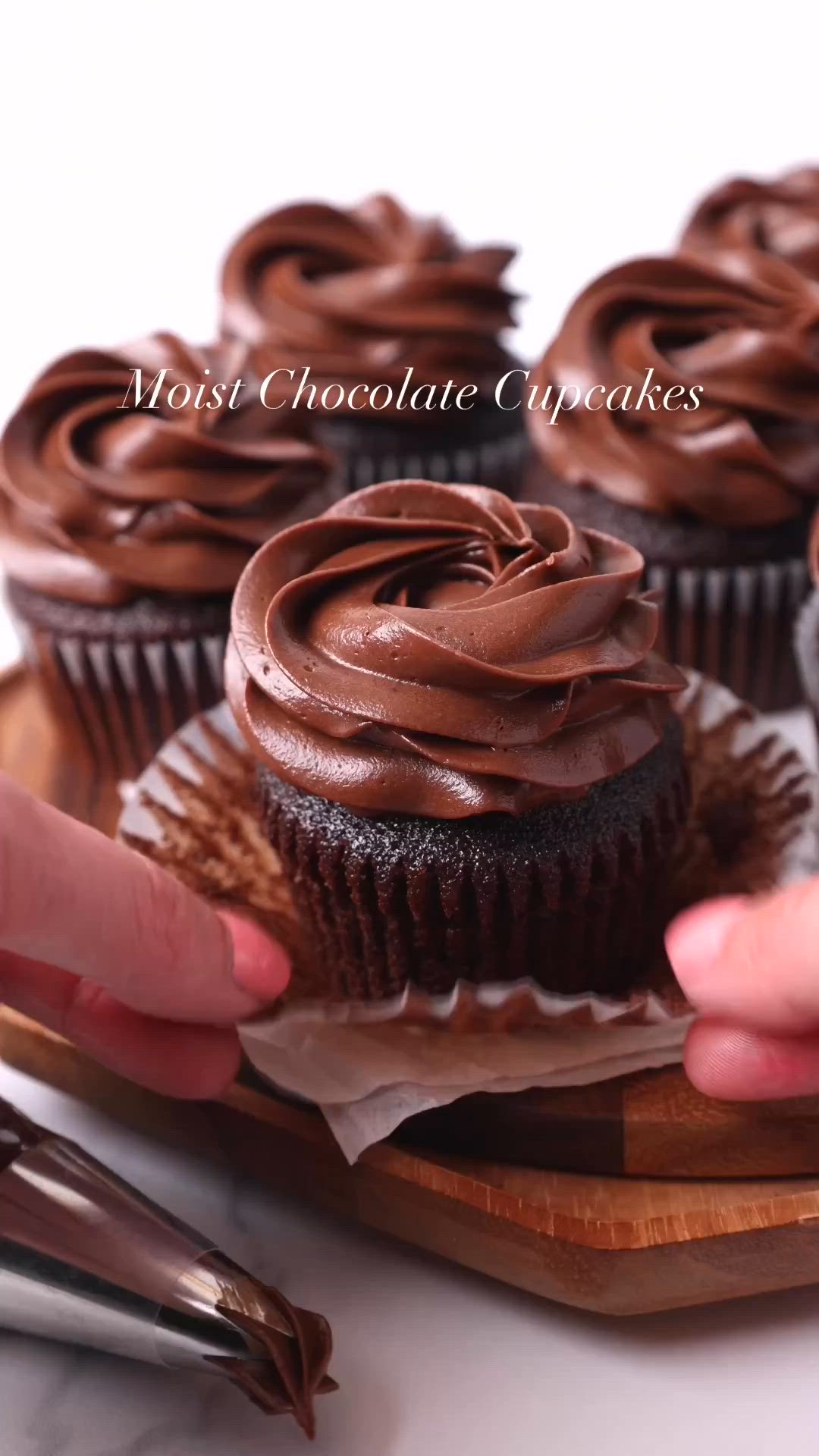 This may contain: chocolate cupcakes with frosting on a wooden tray and two hands reaching for one