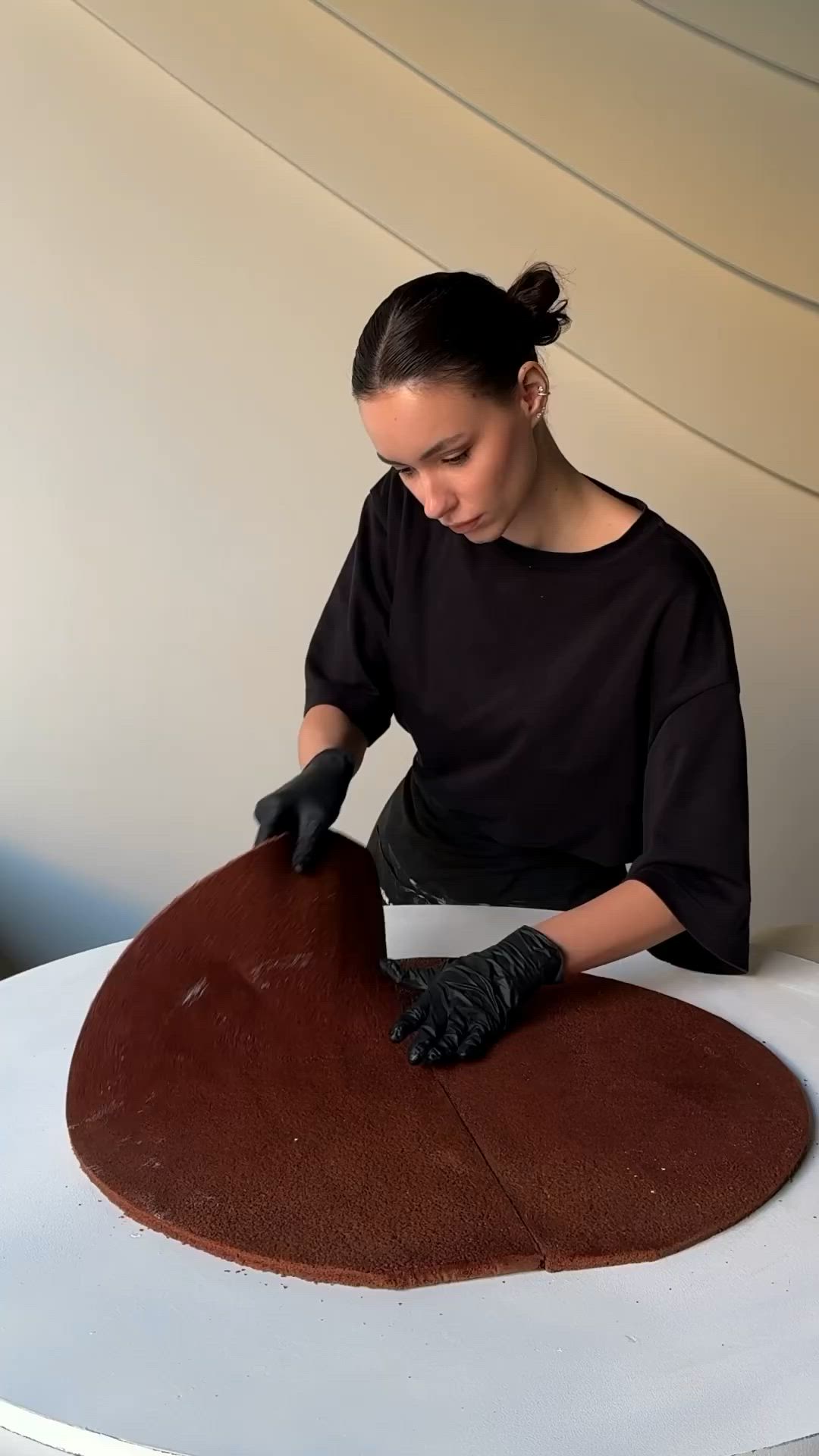 This may contain: a woman in black shirt and gloves making a heart shaped chocolate cake on top of a white table