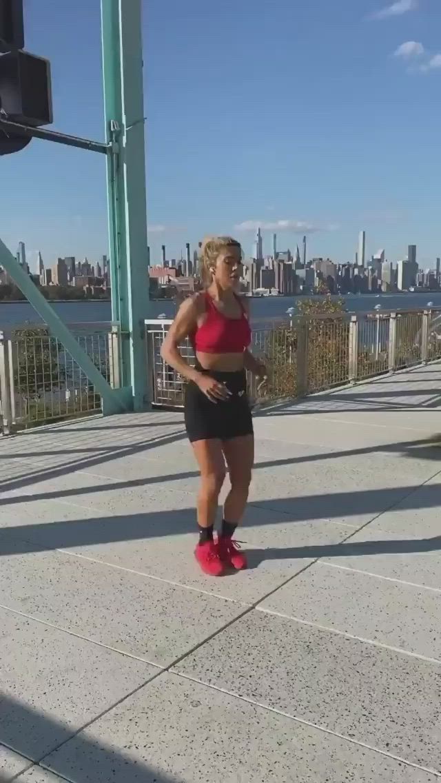 This may contain: a woman in black shorts and red top running across a bridge with the city skyline behind her