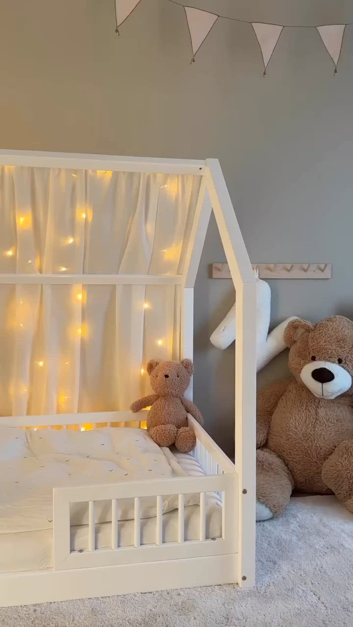 This may contain: a teddy bear sitting in front of a bed with lights on the walls and curtains