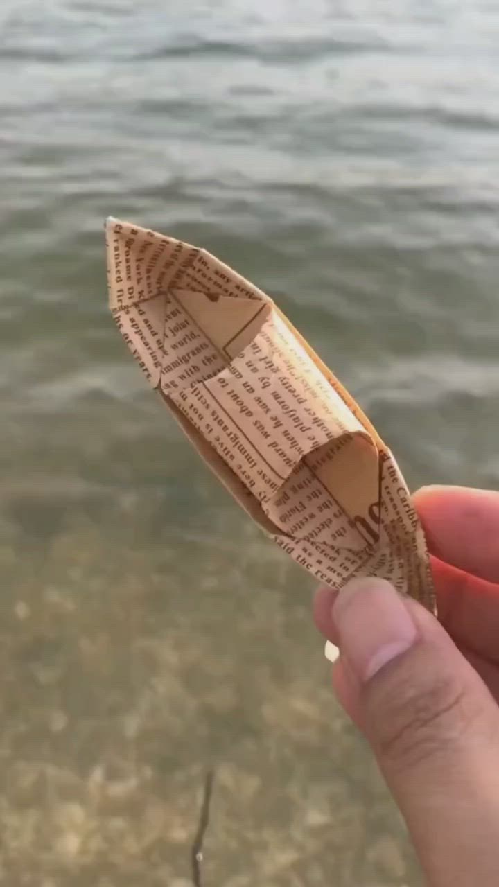 This may contain: someone is holding an origami boat in their left hand near the water's edge