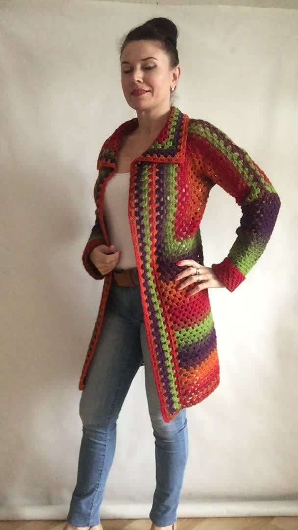 This may contain: a woman standing in front of a white wall wearing a colorful crocheted cardigan