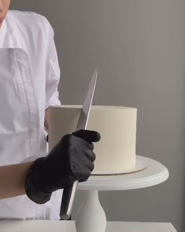 This may contain: a woman in white coat and black gloves decorating a cake with red icing