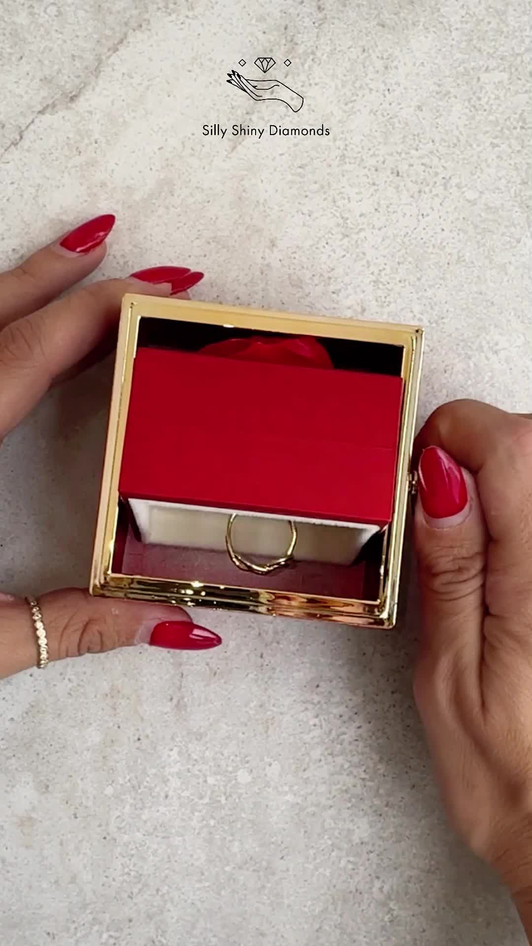 This may contain: a woman's hand with red nails and gold rings on it, next to a white box