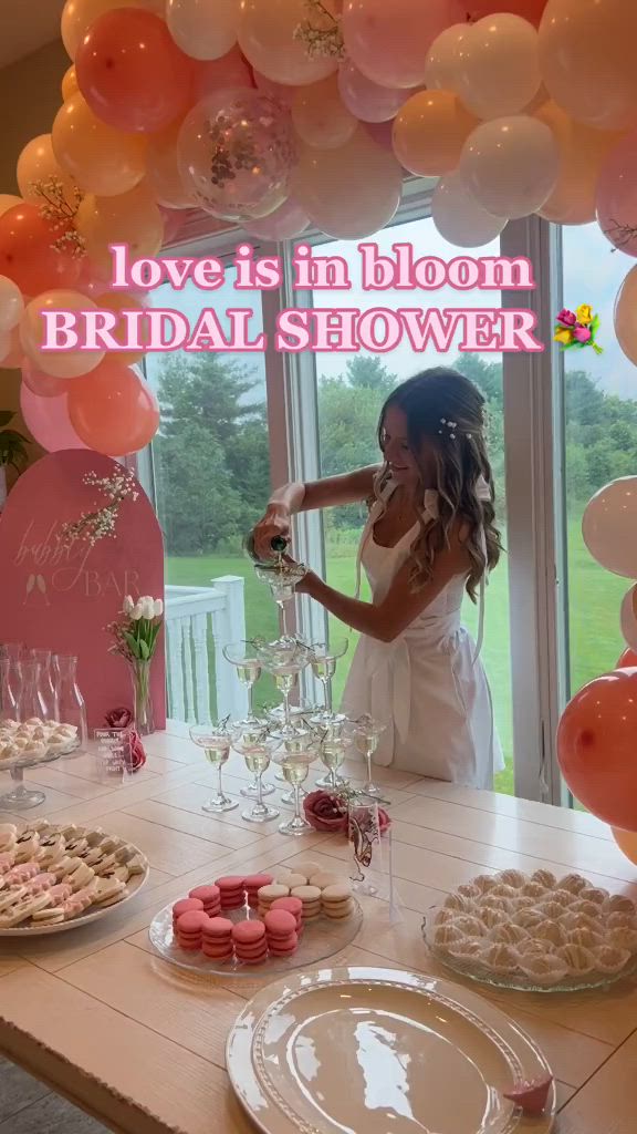 This may contain: a woman standing in front of a table filled with desserts and balloons that read love is in bloom bridal shower