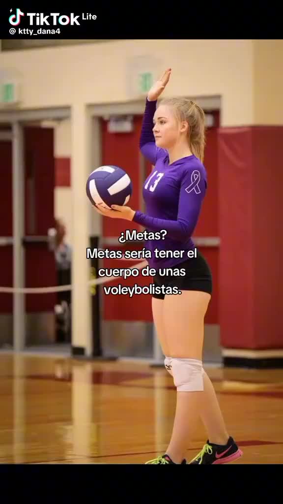 This may contain: a woman in purple shirt and black shorts holding a volleyball ball on top of a gym floor