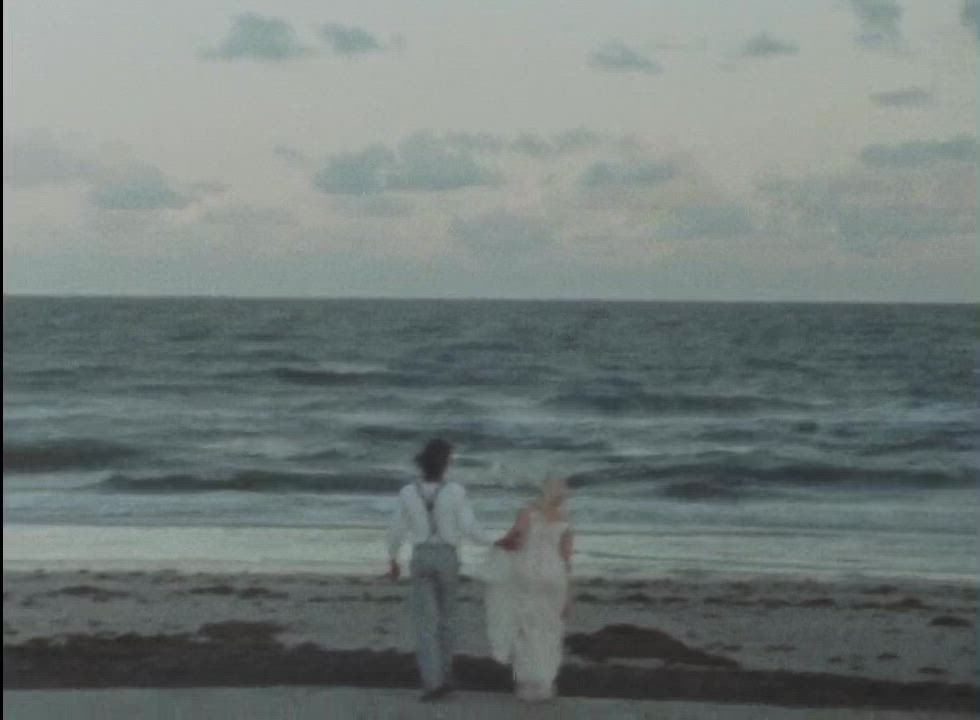 This may contain: a man and woman are walking on the beach with an ocean in the back ground