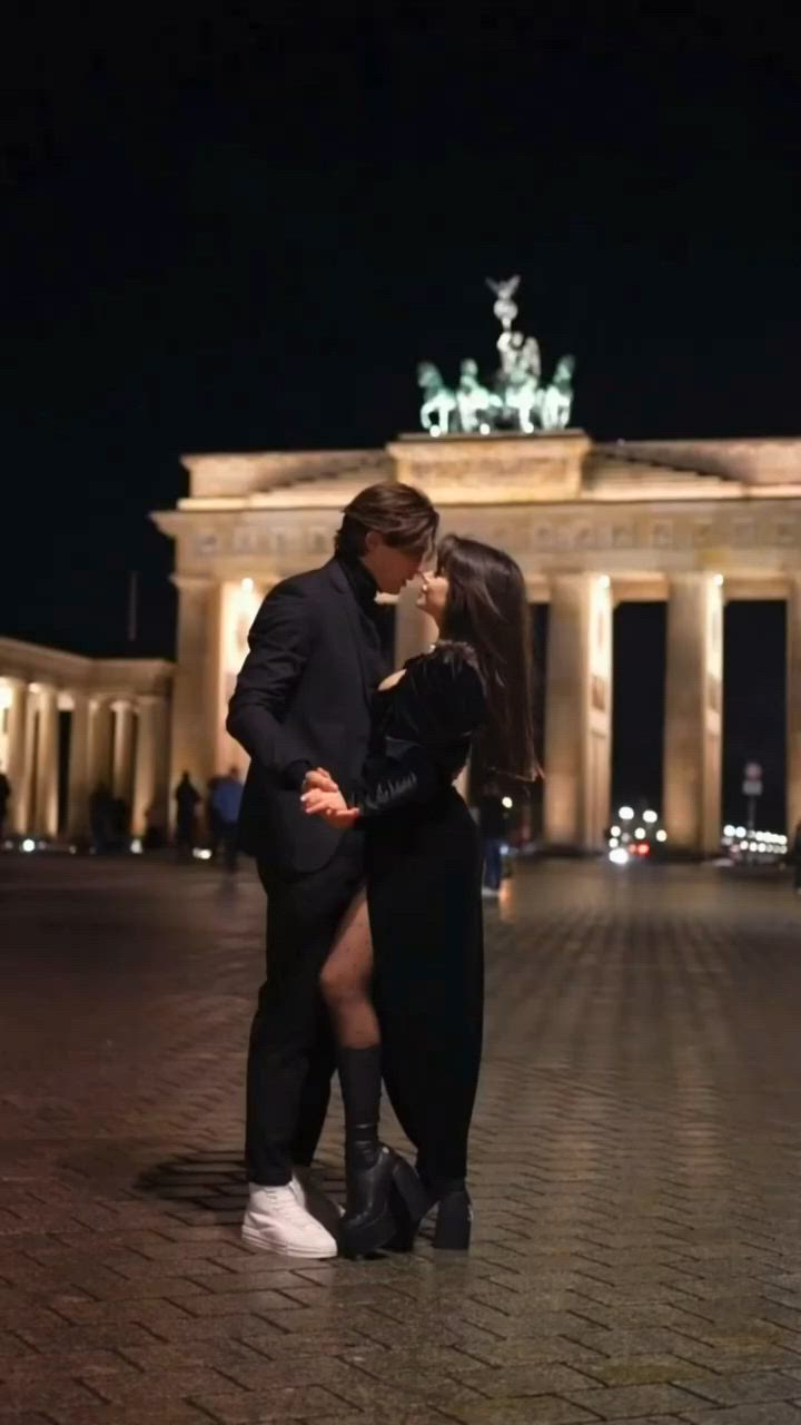 This may contain: a man and woman standing next to each other in front of a building at night