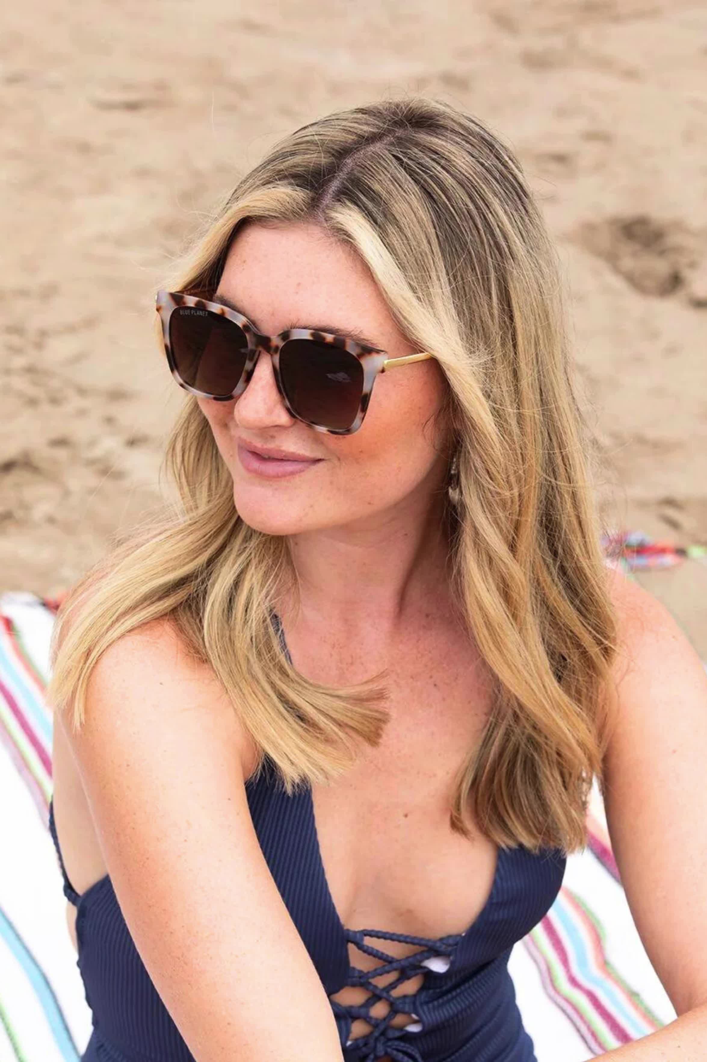 This contains: Woman with blonde hair wearing tortoiseshell sunglasses and a navy blue swimsuit, sitting on a striped beach towel on the sand.