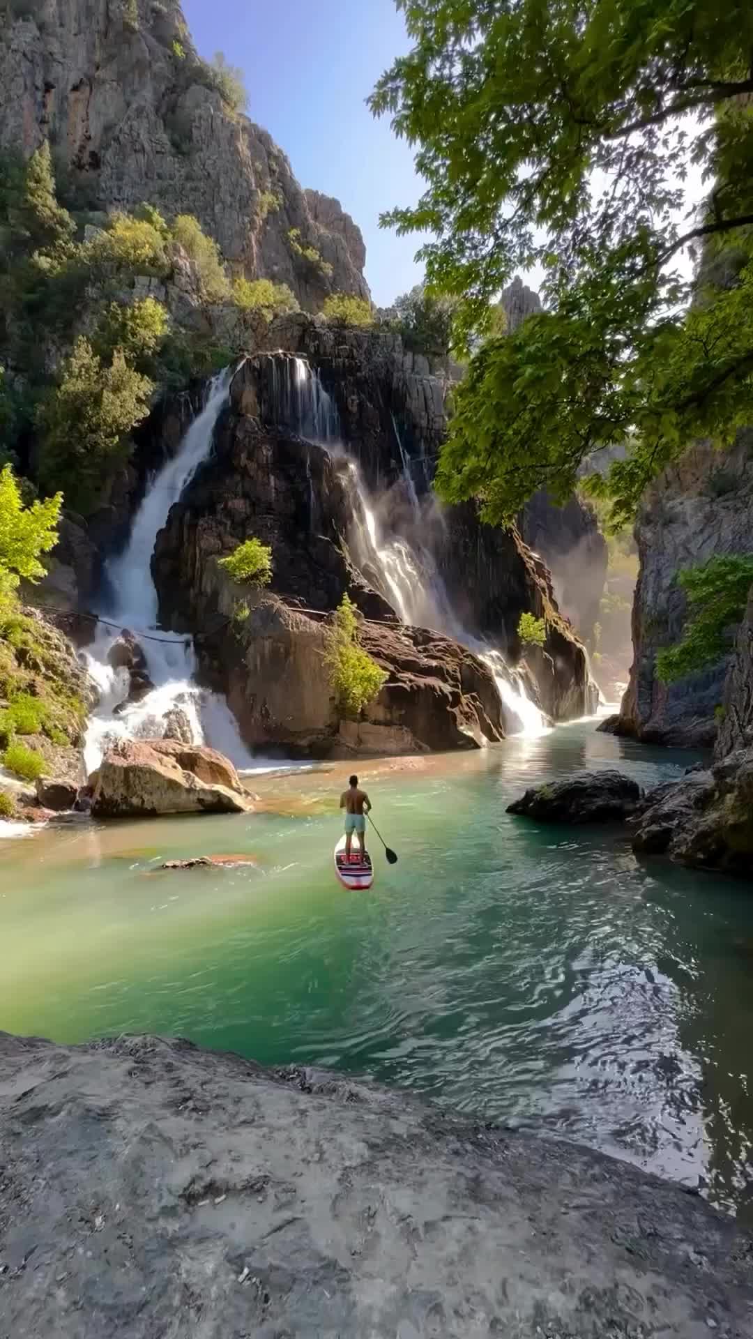 This may contain: a person riding a paddle board in the water near a waterfall and rock face wall