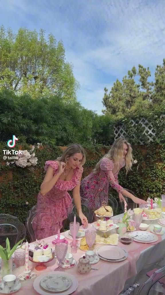 This may contain: two women in pink dresses sitting at a table with plates and cups on it, eating cake