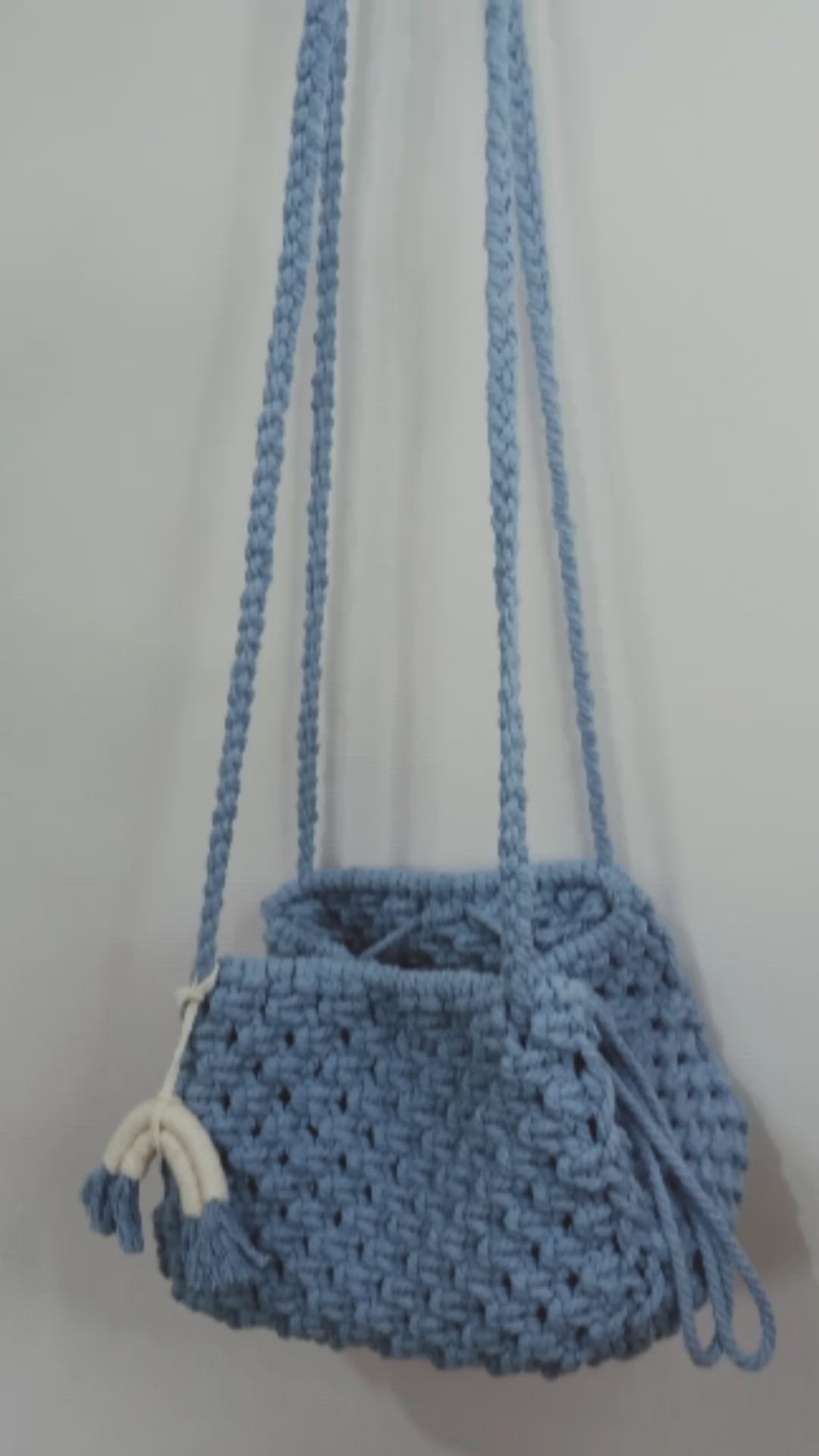 This may contain: a blue crocheted purse hanging from a hook on a wall with two strings