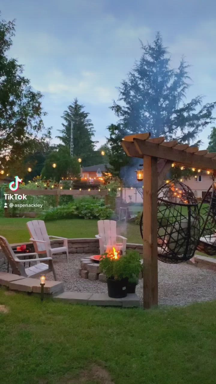 This may contain: an outdoor fire pit surrounded by lawn chairs and lights in the evening with a gazebo behind it