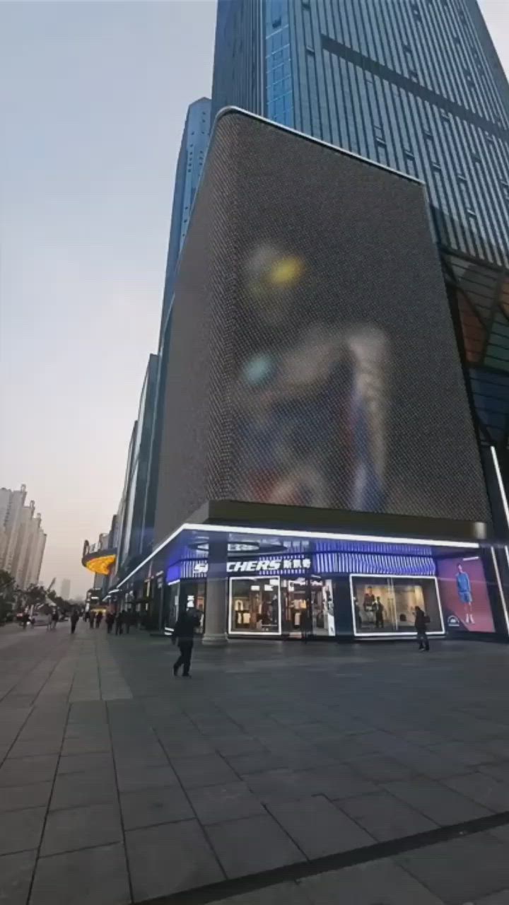 This may contain: people are walking around in front of a tall building that has a giant screen on it