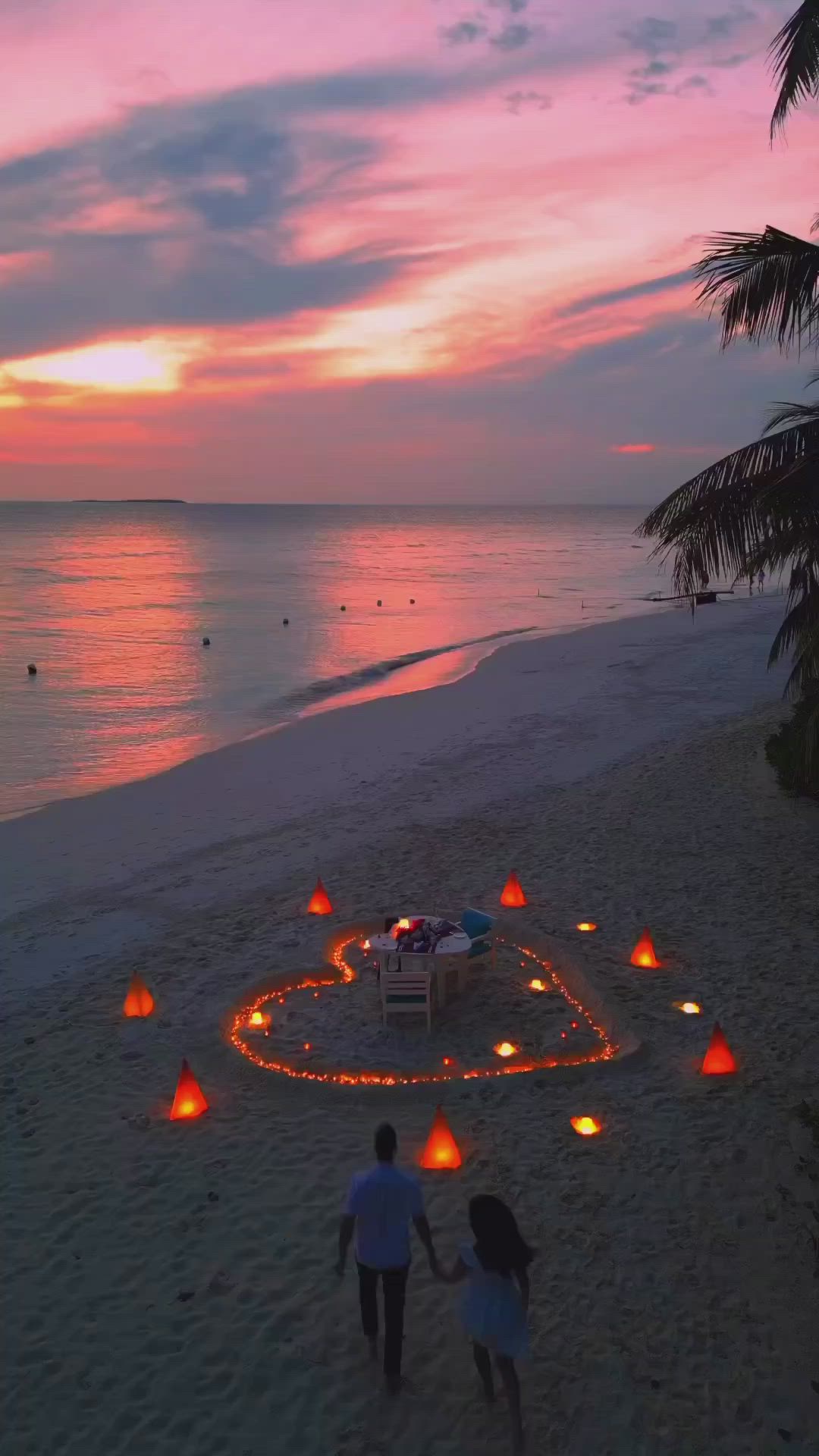 This may contain: two people are walking on the beach with candles in the shape of a heart at sunset