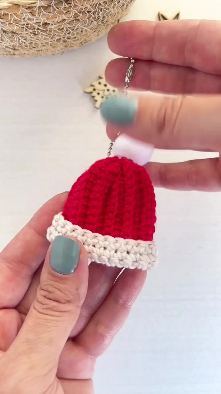 This may contain: a hand holding a crocheted red and white christmas hat ornament on it's finger