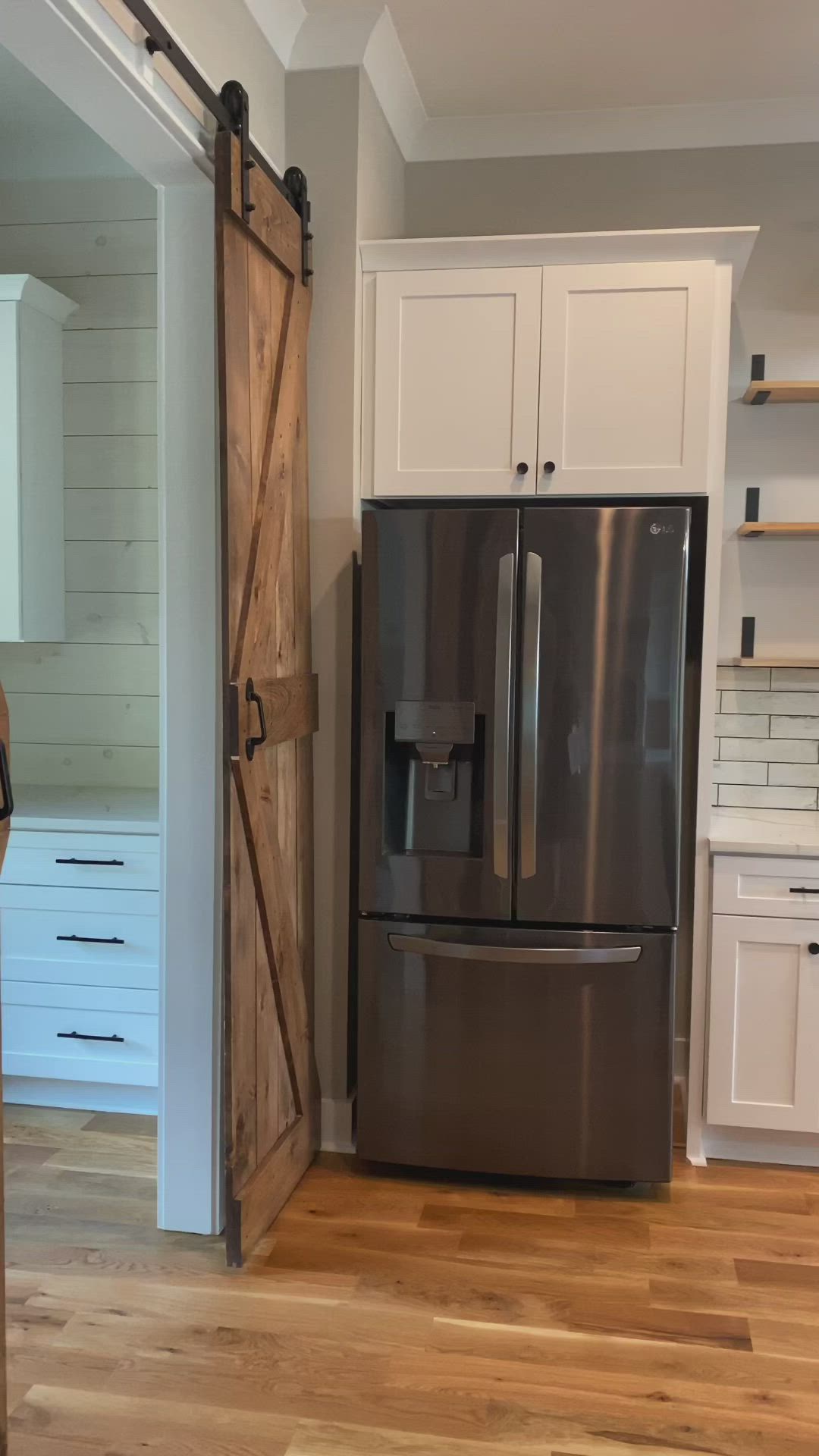 This may contain: a stainless steel refrigerator in a kitchen with white cabinets and wood flooring that has sliding barn doors