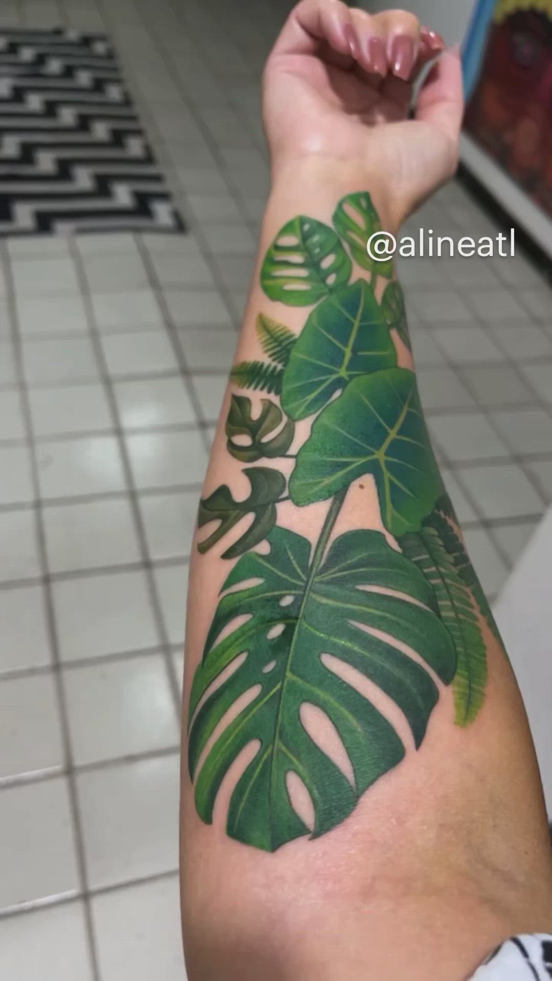 This may contain: a person with a green leaf tattoo on their arm and hand, in front of a tiled floor