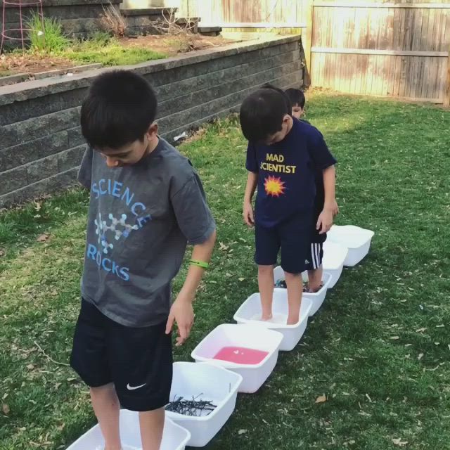 This may contain: three young boys playing with water and sand in plastic containers on the grass near a brick wall