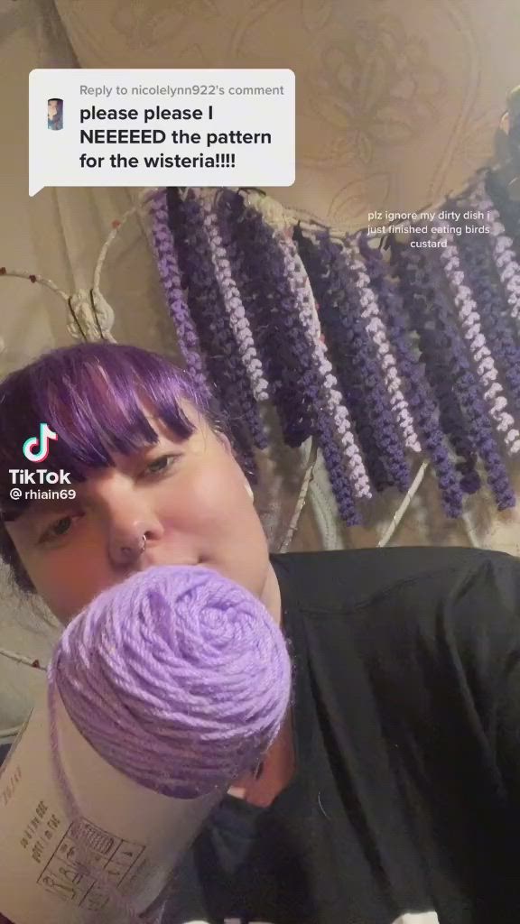 This may contain: a woman with purple hair holding a ball of yarn in front of her face and looking at the camera