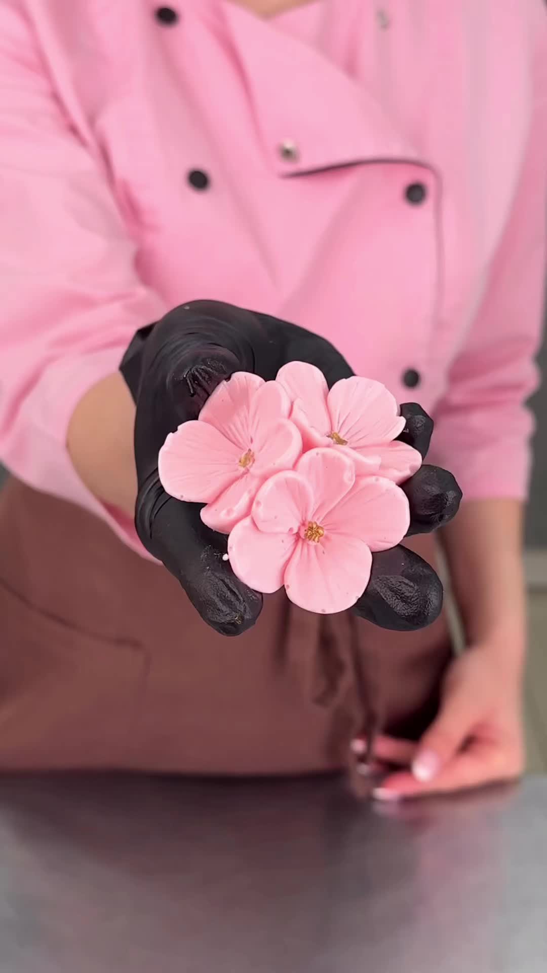 This may contain: a woman in pink shirt and black gloves holding up a flower with one hand on top of the other