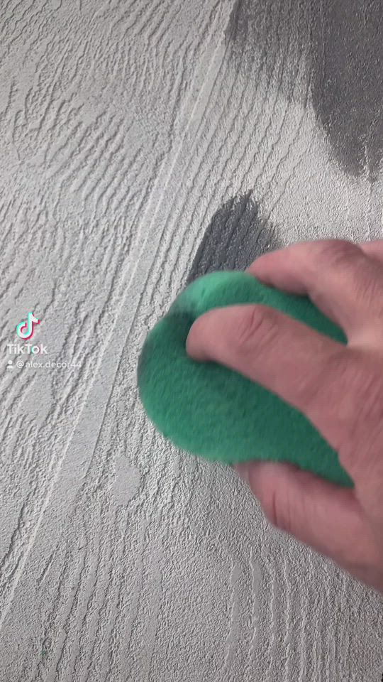 This may contain: a hand holding a green sponge on top of a wooden table next to a gray wall