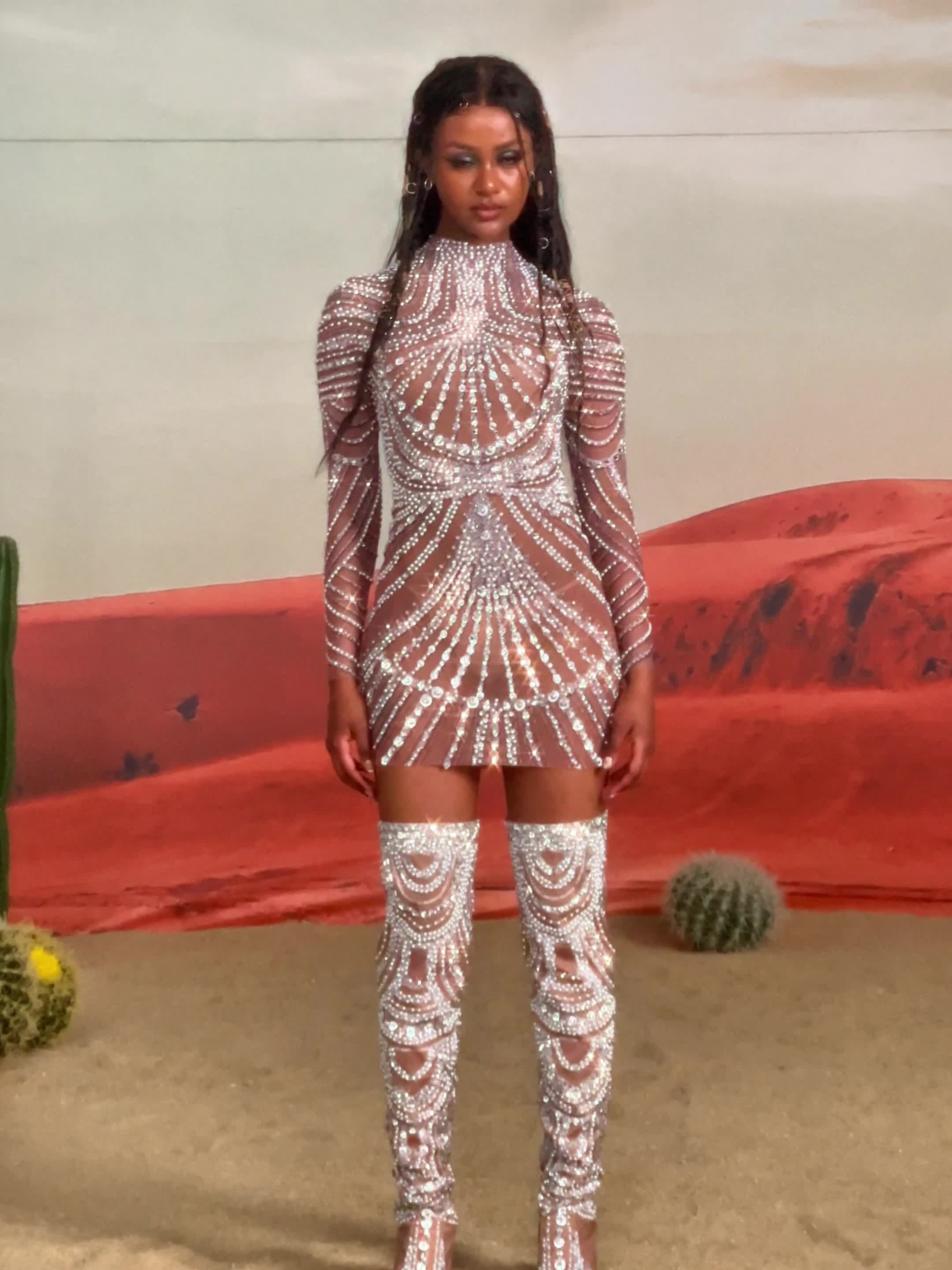 This may contain: a woman standing in front of a desert backdrop wearing white sequins and thigh high boots