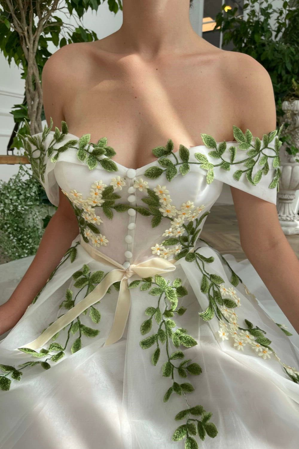 The White tulle off-shoulder corset dress with built-in pads and boning is cinched at the waist and Tea-length style. The Pleated skirt has side pockets. It is Embellished with embroidered green leaves and flowery details. Decorative white buttons and ivory ribbon belt.