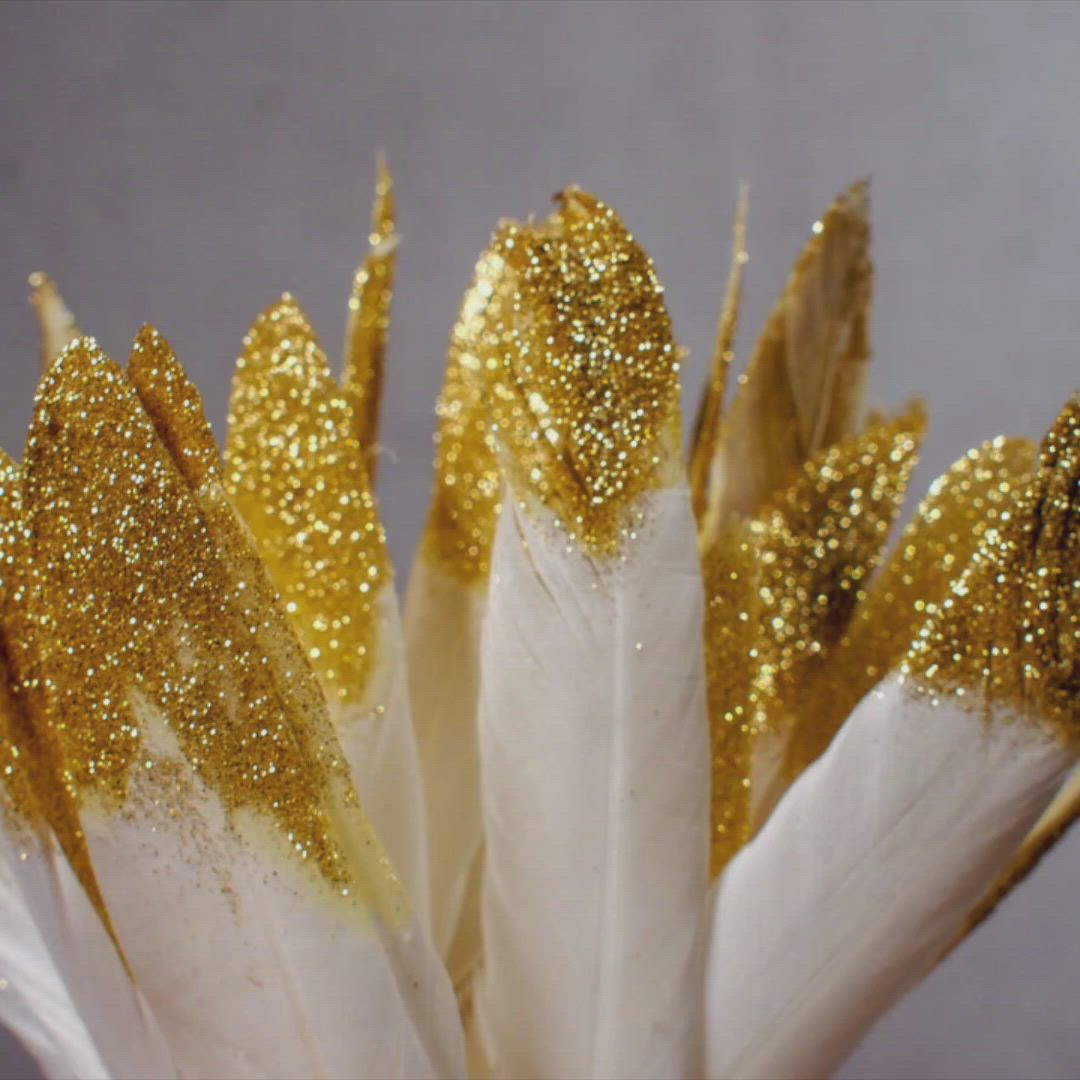 This may contain: gold and white feathers with some glitter on them are in a vase or flower pot