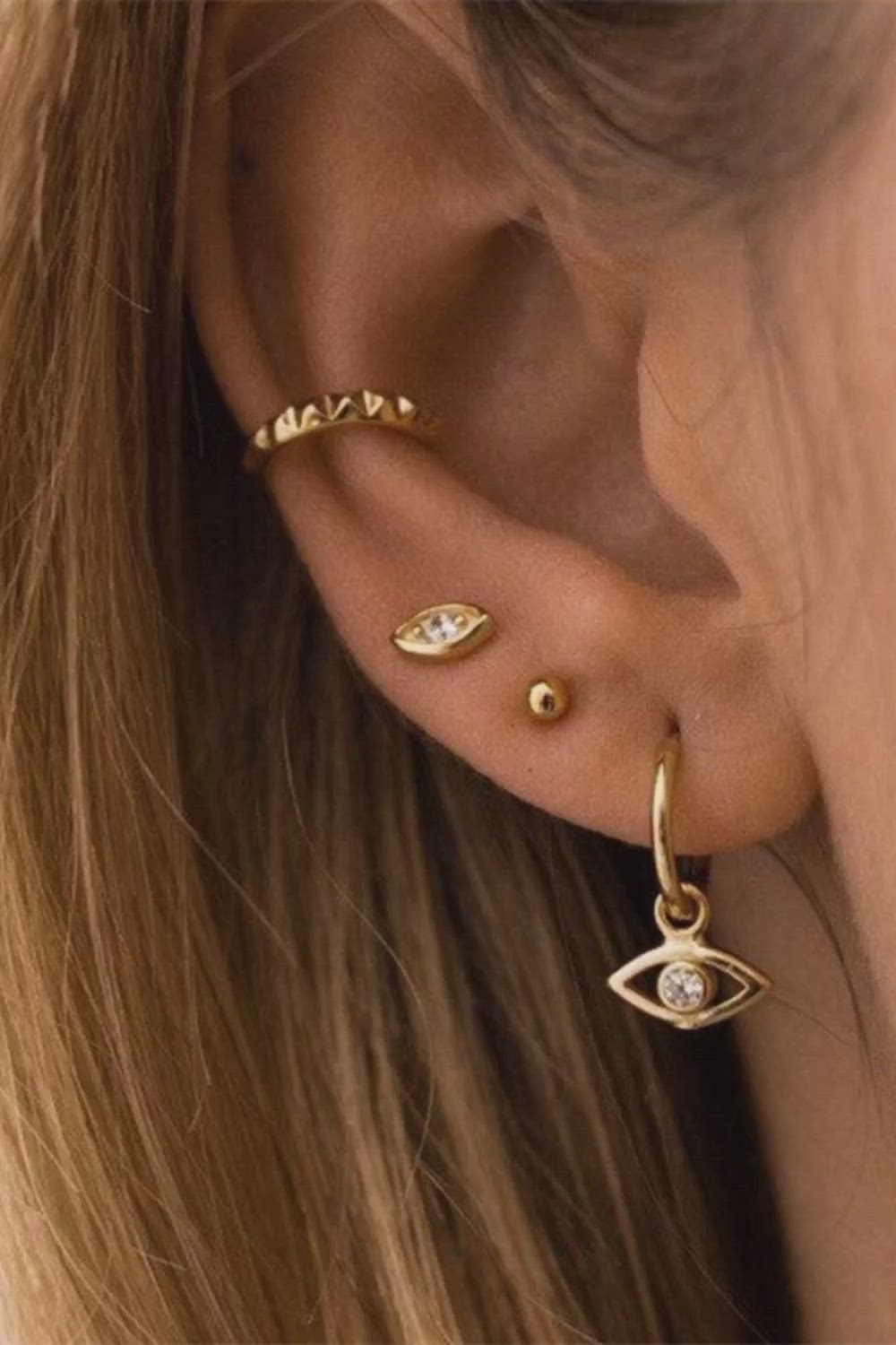 This may contain: a woman wearing gold ear piercings with an evil eye on the top and diamond eyes on