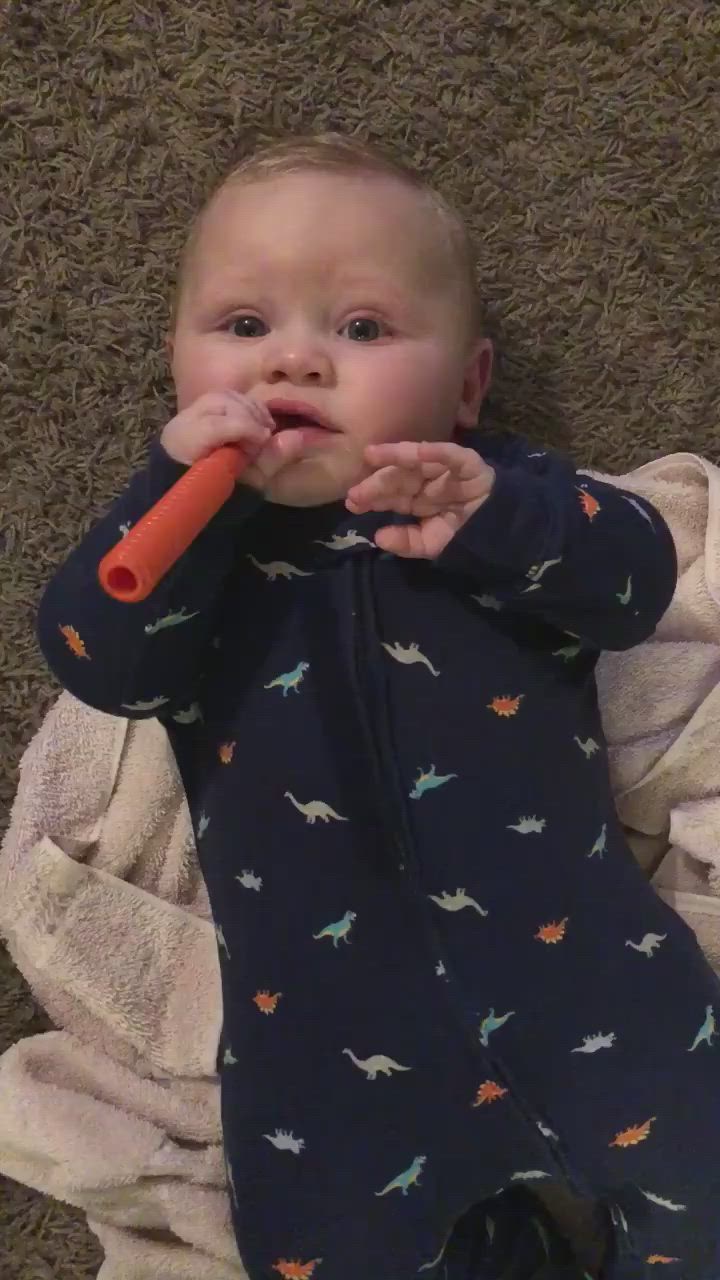 This may contain: a baby laying on the floor chewing on an orange toy while holding it in its mouth