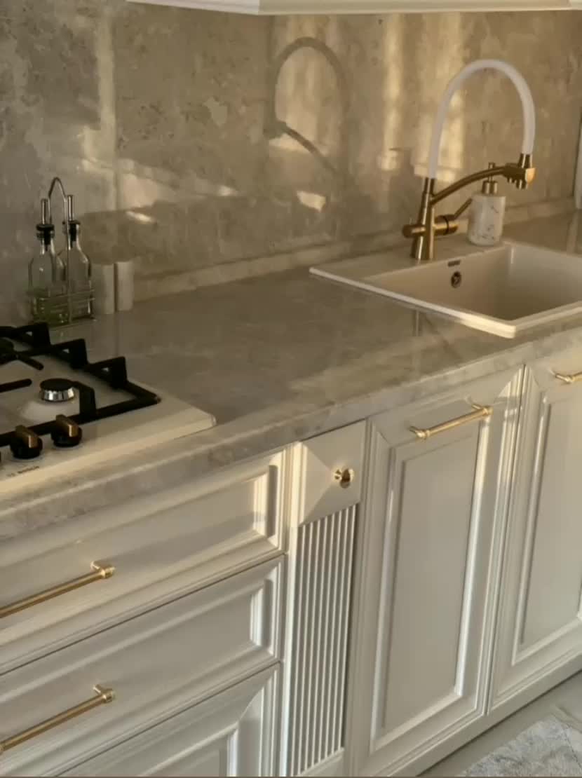 This may contain: a white kitchen with marble counter tops and gold trim on the sink, stove top and cabinets