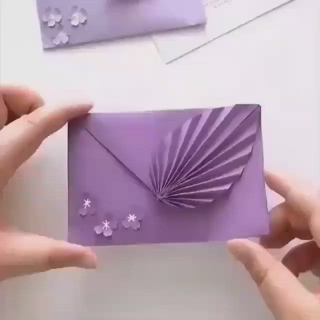 This may contain: two hands holding a purple card with white flowers on it and an envelope in the middle