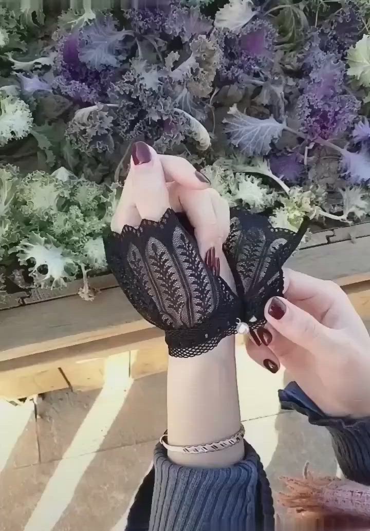 This may contain: a woman wearing black lace gloves with flowers in the background and her hands holding onto an object